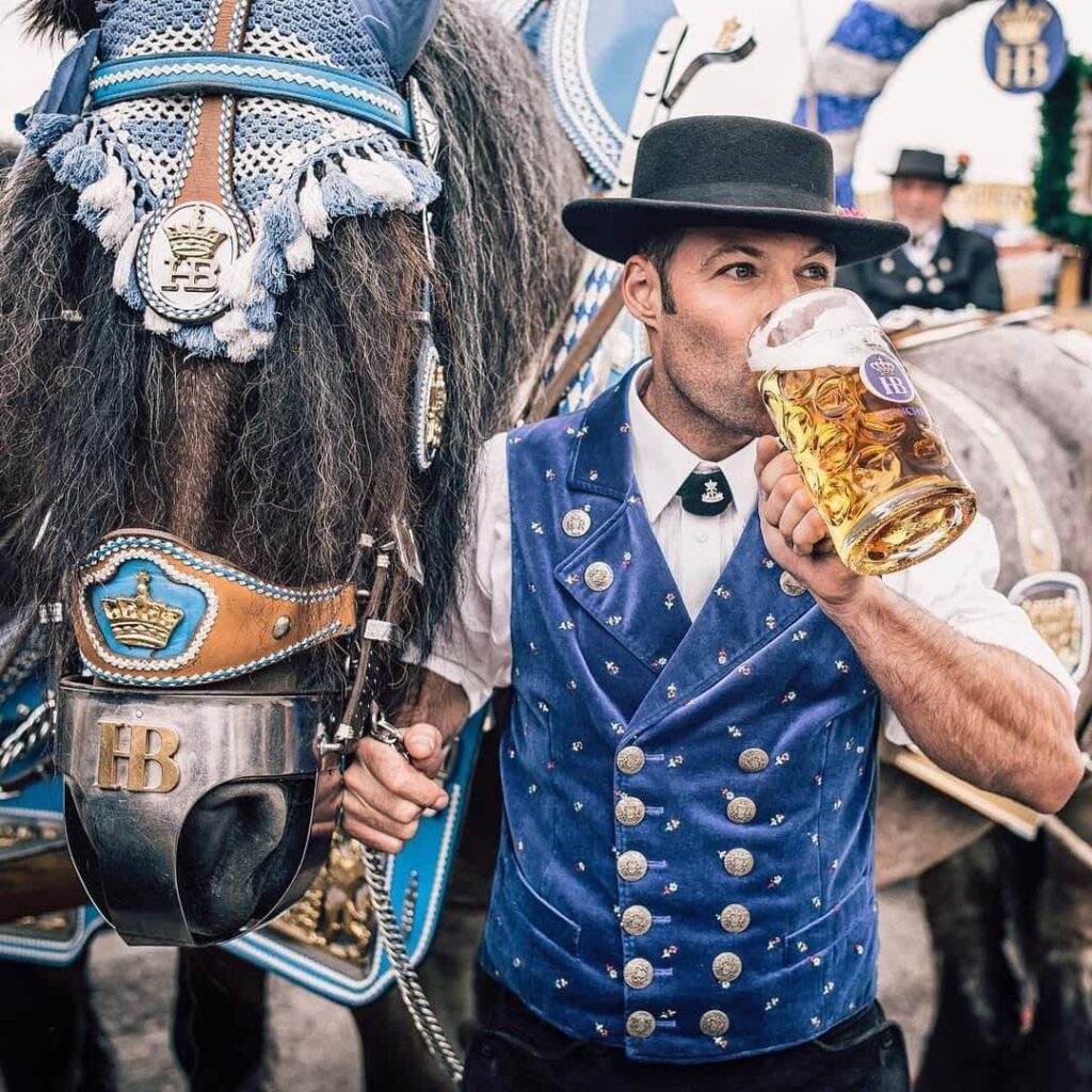 Man in traditional German attire drinking a stein of beer in front of a horse.